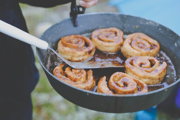 Expedition Fry-Bake pan filled with golden-brown cinnamon rolls cooking outdoors, showing the versatile baking capabilities of this durable aluminum camping cookware. Close-up view of spiral pastries in black pan against blurred natural background.