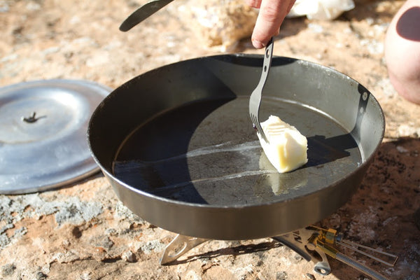 Expedition Fry-Bake pan shown outdoors with butter being added to its dark-coated cooking surface. The 10.5-inch aluminum pan sits on a natural surface with its lid visible nearby.