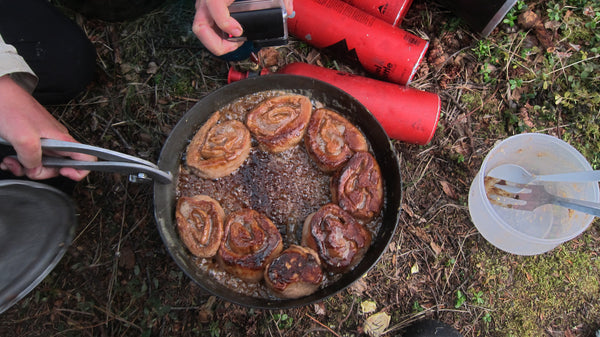 Expedition Fry-Bake pan showing freshly baked cinnamon rolls cooking on a campsite ground, with red thermoses and camping utensils visible around the pan, demonstrating outdoor baking capabilities.