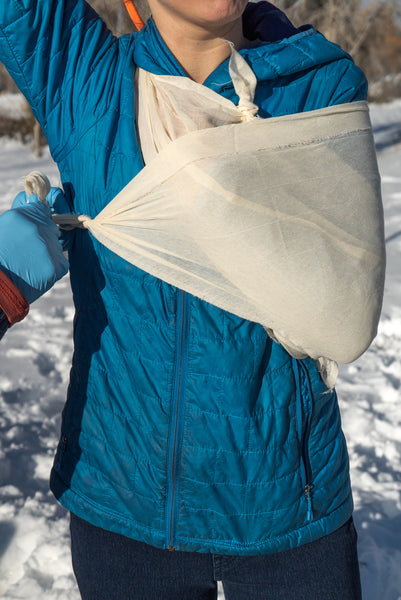Cravat (Triangle Bandage) demonstrated as an arm sling on person wearing blue winter jacket in snowy outdoor setting, showcasing practical first aid application of muslin bandage