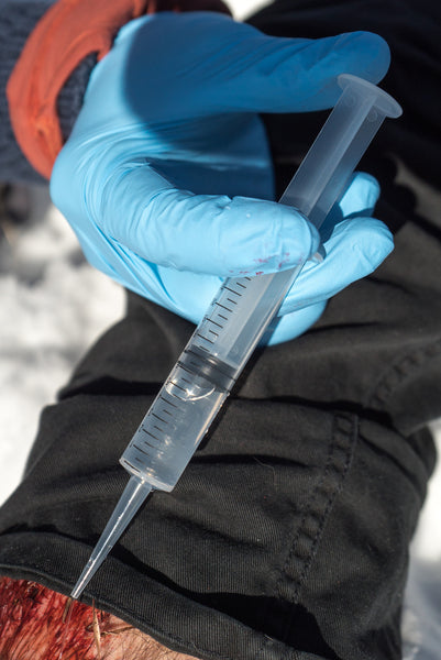 Irrigation Syringe 12cc held in blue-gloved hand, demonstrating clear plastic medical syringe with measurement markings for precise wound cleaning and debris removal in wilderness first aid situations