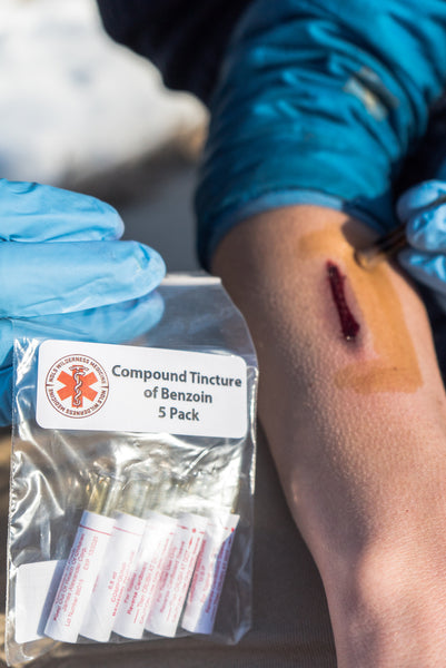 Benzoin Tincture 5-pack shown with medical gloves treating a wound, demonstrating its use in wilderness first aid. Package displays medical symbol and contains single-use applicator vials for wound care and blister management.