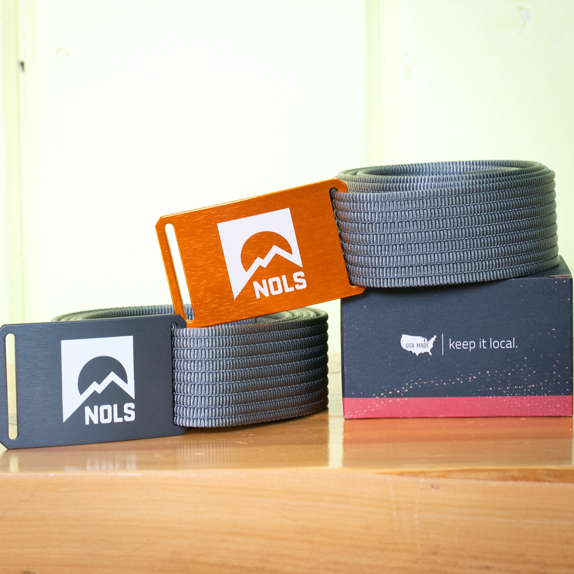 GRIP6 Belt with NOLS Logo featuring gunmetal and orange aluminum buckles with gray nylon webbing straps, displayed alongside product packaging showing Keep it Local slogan and NOLS mountain logo design